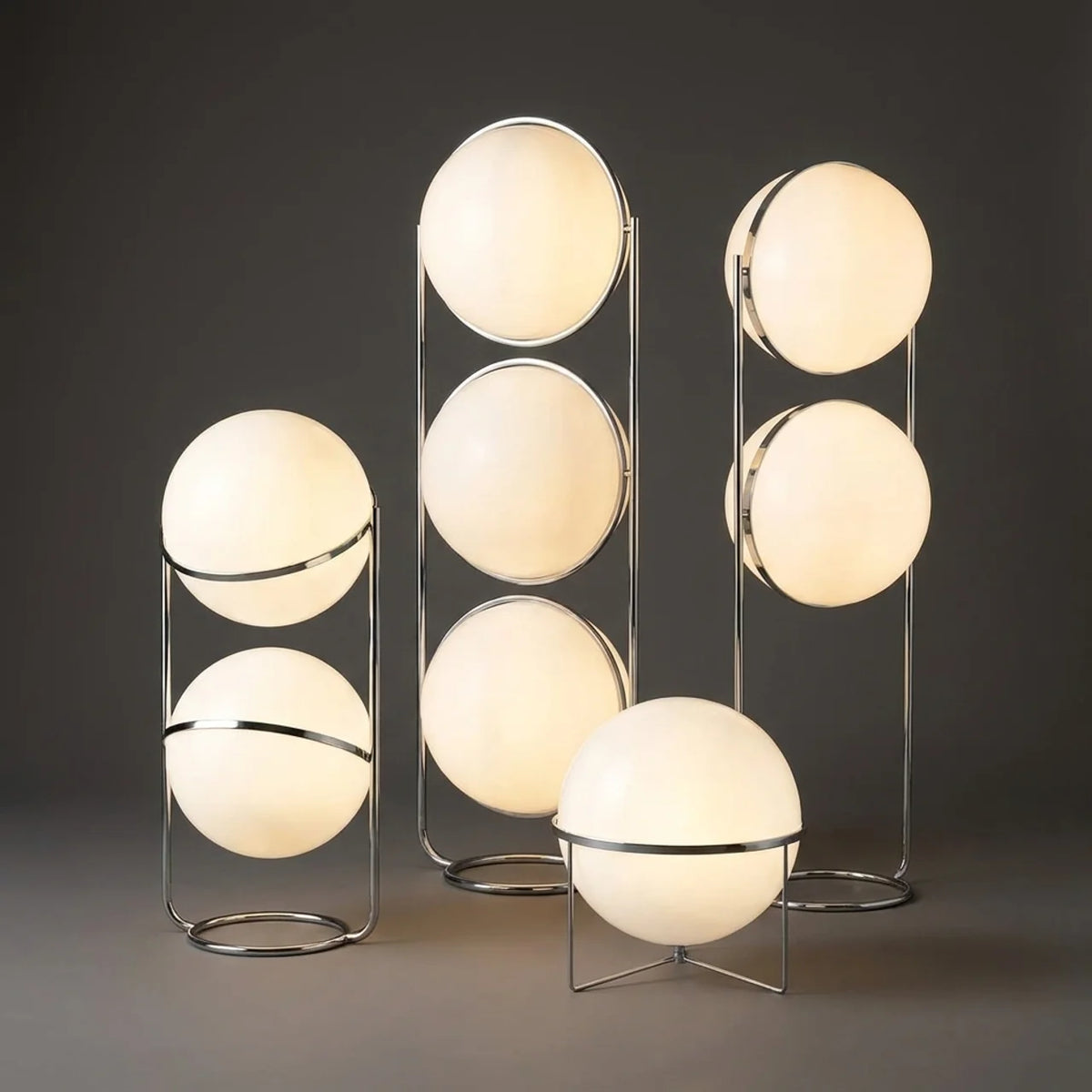Three modern floor lamps with glowing oval frosted glass globes and slim metallic stands, arranged on a dark gray studio background.