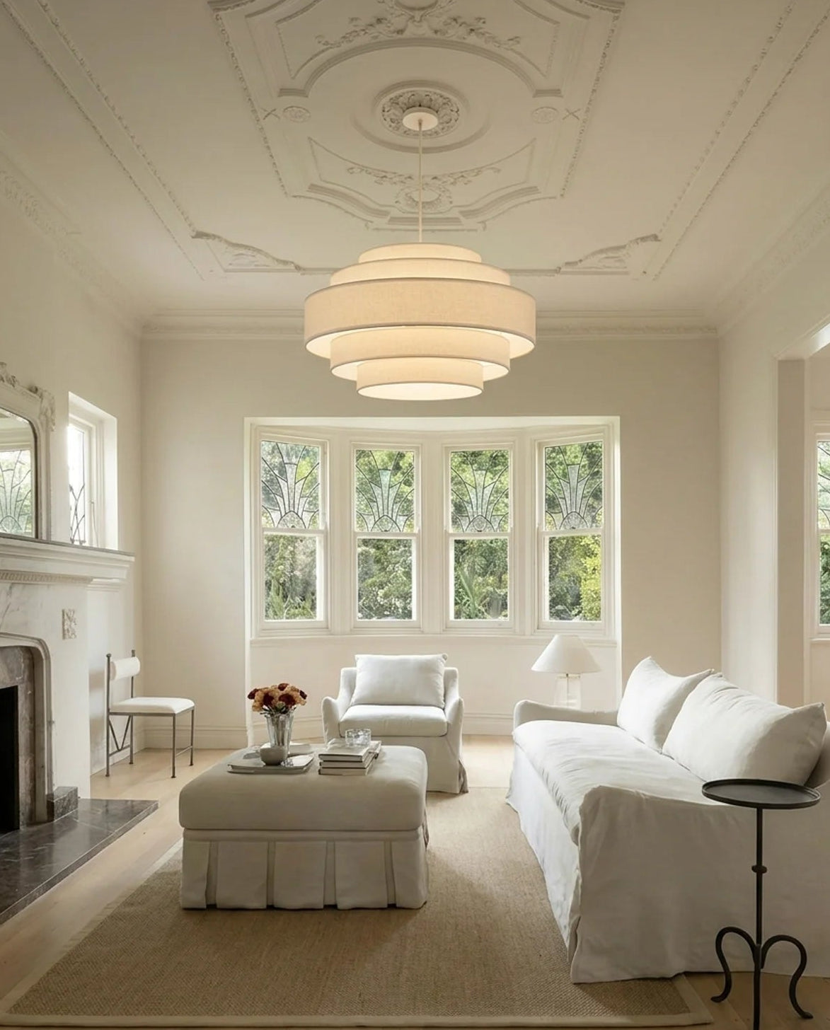 A bright, elegant living room featuring a large tiered white pendant light hanging from a decorative white ceiling. The room is furnished with a white slipcovered sofa, matching armchair, and a central ottoman on a beige rug, with a fireplace on the left and tall windows in the back showing green foliage outside.