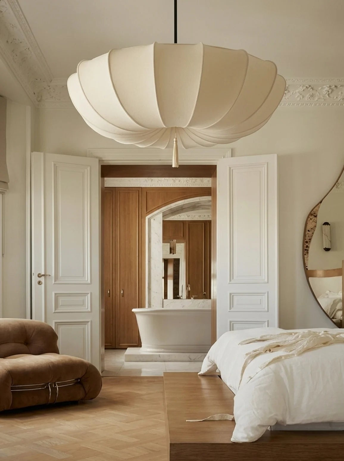A large cream-colored paper lantern pendant light hangs in a beautifully styled, warm-toned bedroom with a wooden platform bed, white bedding, and a brown leather lounge chair.
