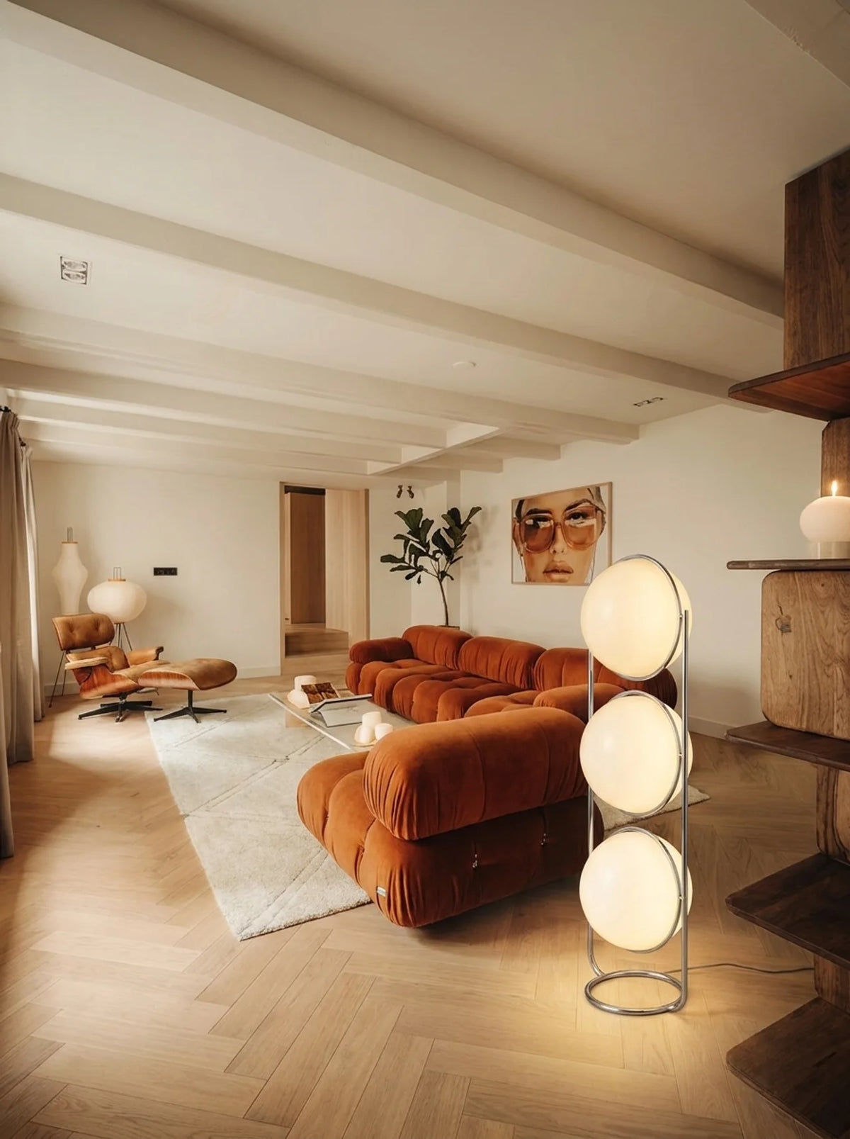 A modern living room with a prominent terracotta orange modular sofa, a white area rug, light wood herringbone flooring, and a tall three-light floor lamp in the foreground.