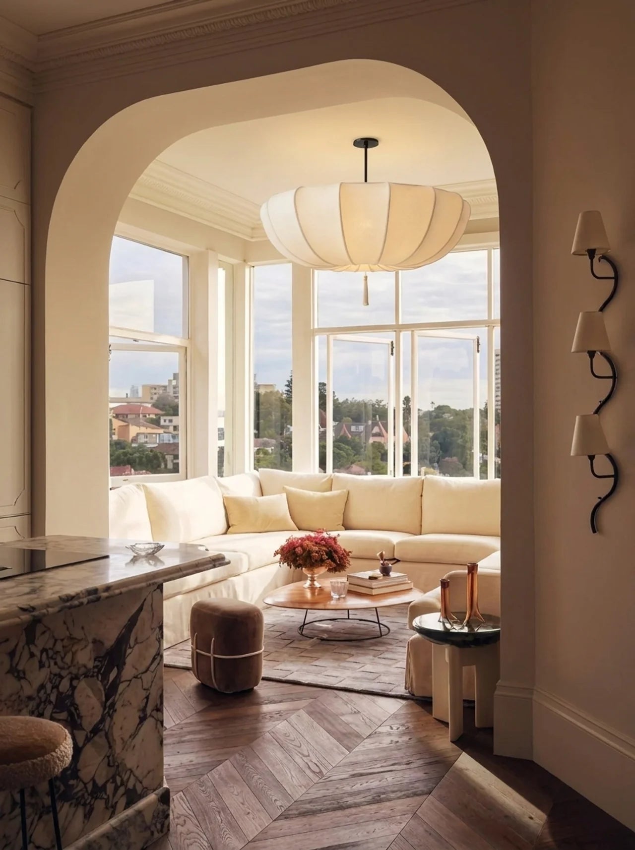 A modern living room interior with a large cream sectional sofa, a white paper lantern-style pendant light, wood herringbone flooring, and an arched doorway leading to a sunlit balcony area.