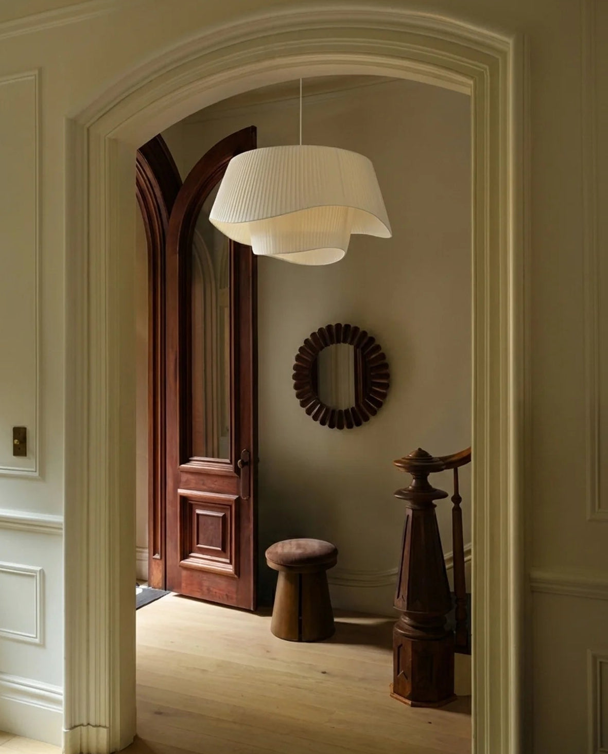 A modern pleated white pendant light hangs in a classic home entryway, viewed through an arched doorway. The room features warm wood floors, a dark wood door, a round sunburst mirror, and a wooden stool.