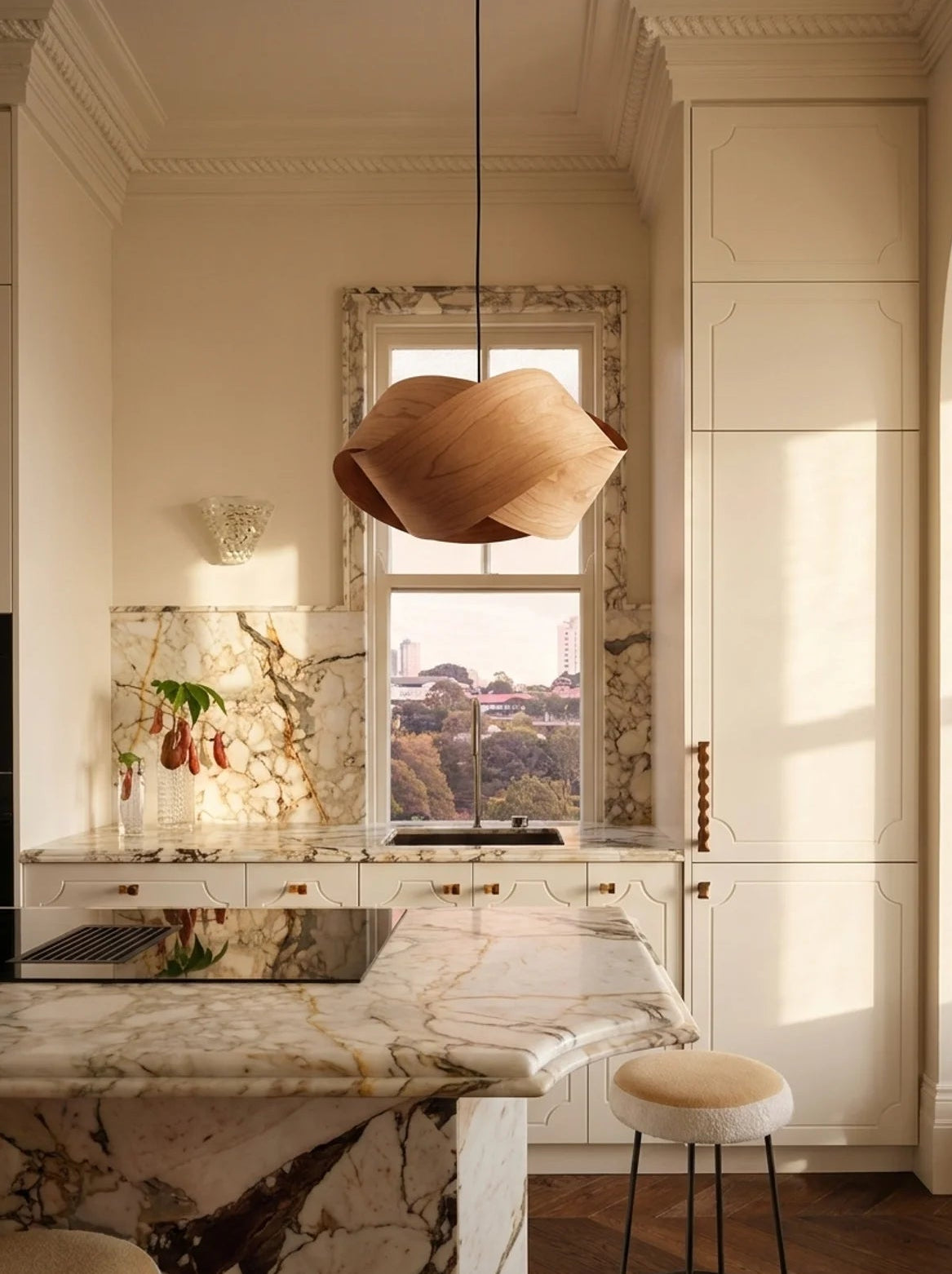 A modern kitchen interior with cream cabinetry, a marble island and backsplash, a large sculptural wooden pendant light, and a window looking out onto a park view.