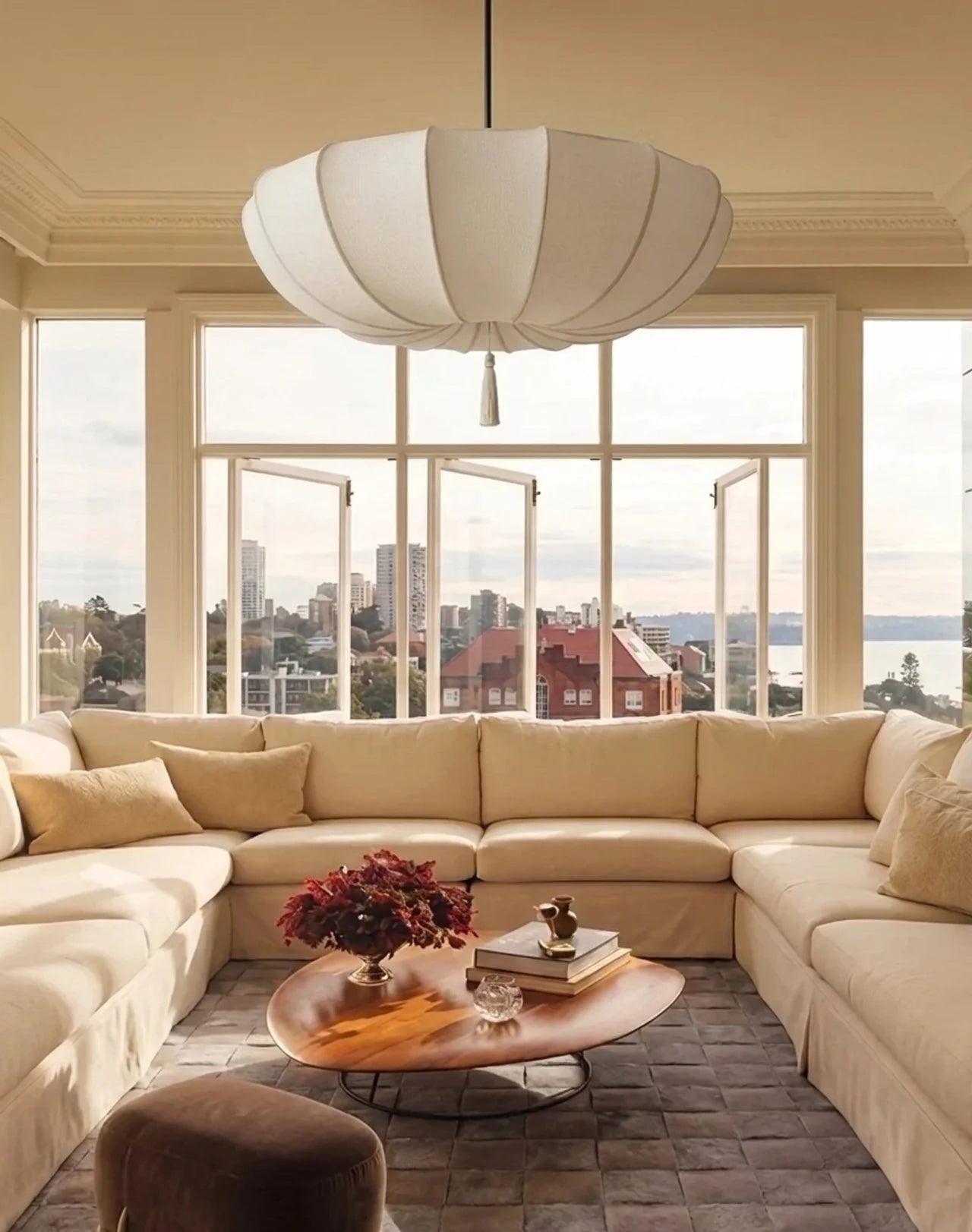 A modern living room featuring a large cream sectional sofa, a white paper lantern-style pendant light, and a round wooden coffee table, set against large windows with a city and ocean view.