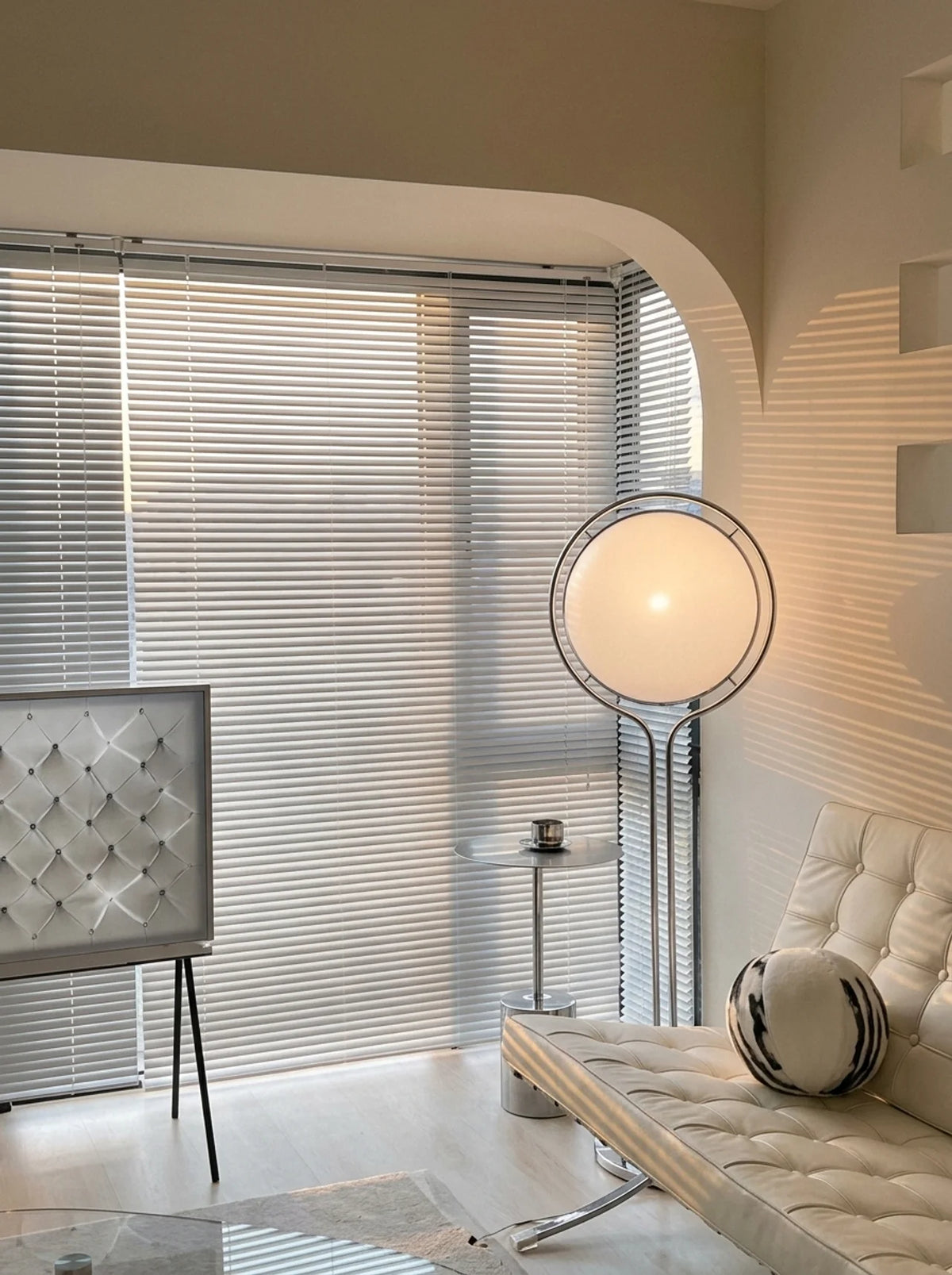 A modern living room corner with white horizontal blinds covering a large window, a round floor lamp with a glowing white globe shade, a tufted cream chaise lounge with a striped accent pillow, and warm sunlight casting striped shadows across the wall.