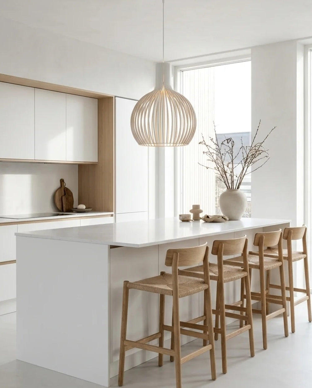 A modern kitchen island with a white countertop and base, paired with four light wood bar stools. A white pendant light hangs above, and the background shows white cabinetry with light wood trim in a bright, minimalist kitchen.