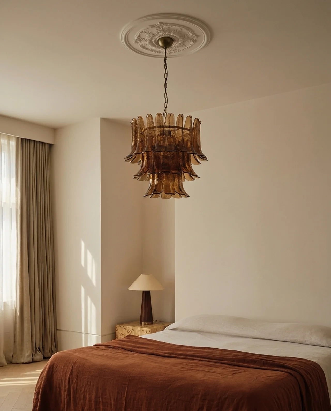 An amber-brown glass chandelier with a tiered, fluted design hangs from a decorative ceiling medallion in a minimalist, warm-toned bedroom.