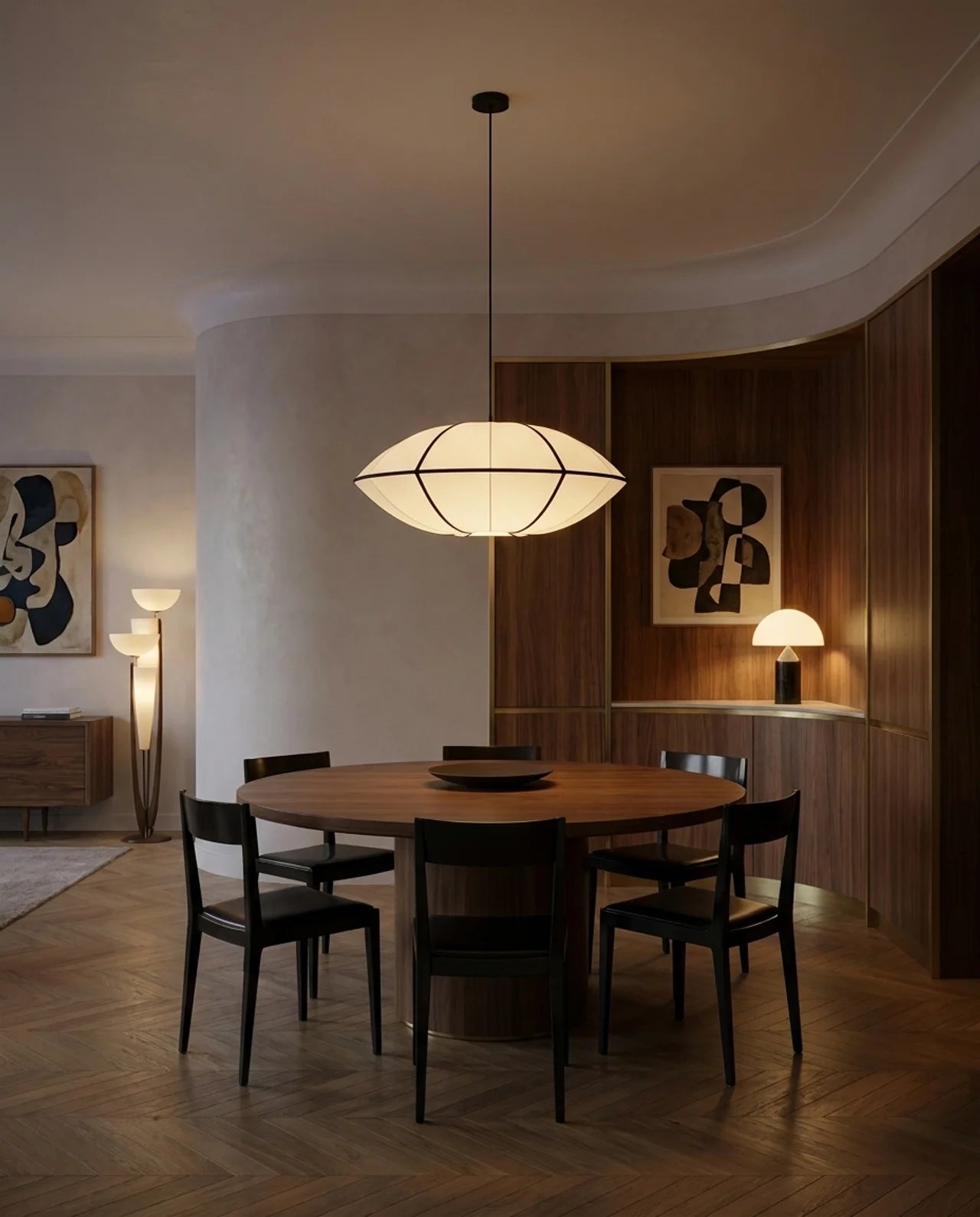 A modern dining room scene featuring a round wooden dining table surrounded by six black chairs under a large, geometric, dome-shaped pendant light. The room has warm wood paneling on the right, textured off-white walls on the left, and herringbone wood flooring.