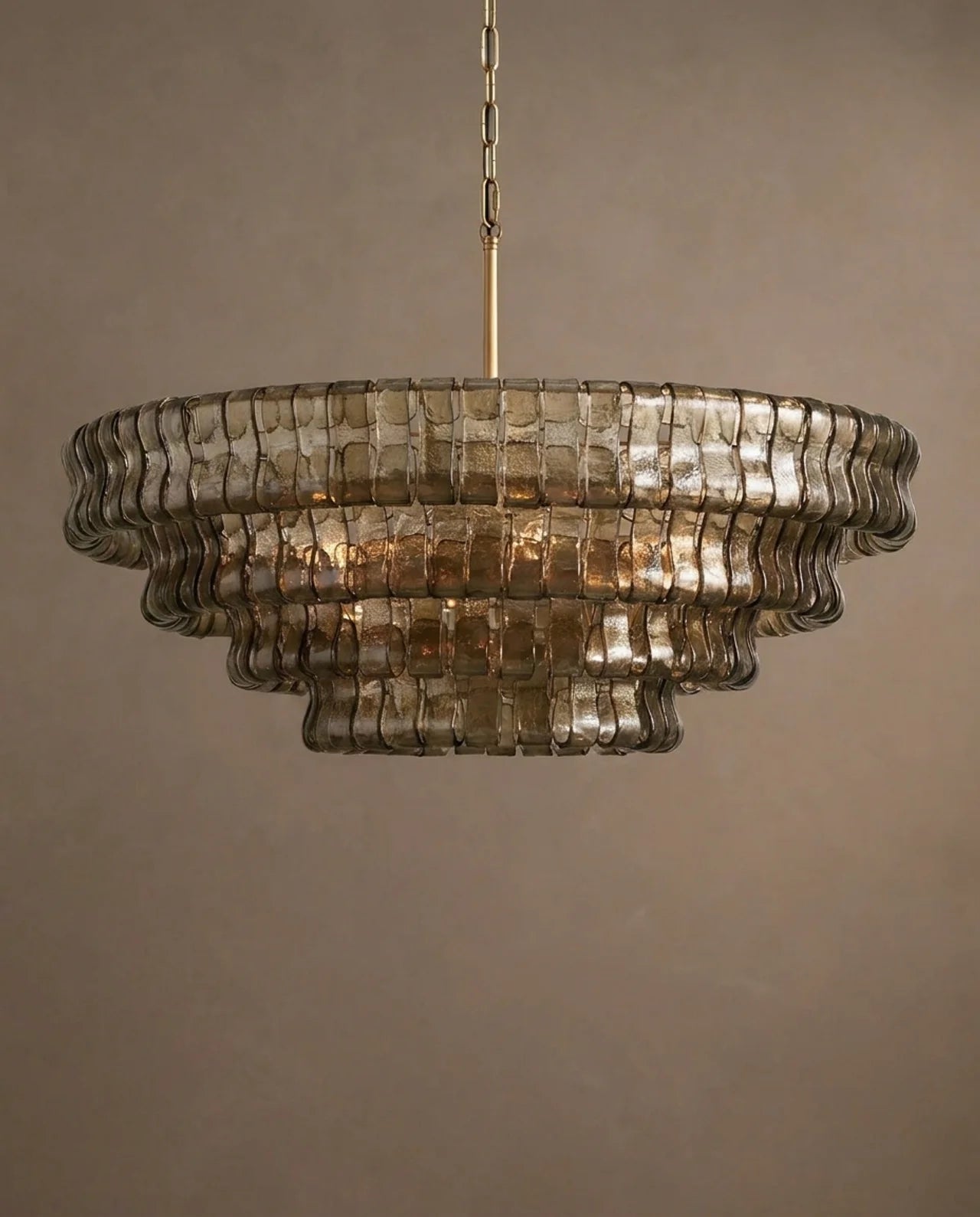 A tiered chandelier with textured, wavy smoked glass shades and a gold-toned hanging chain, shown against a neutral warm gray background.