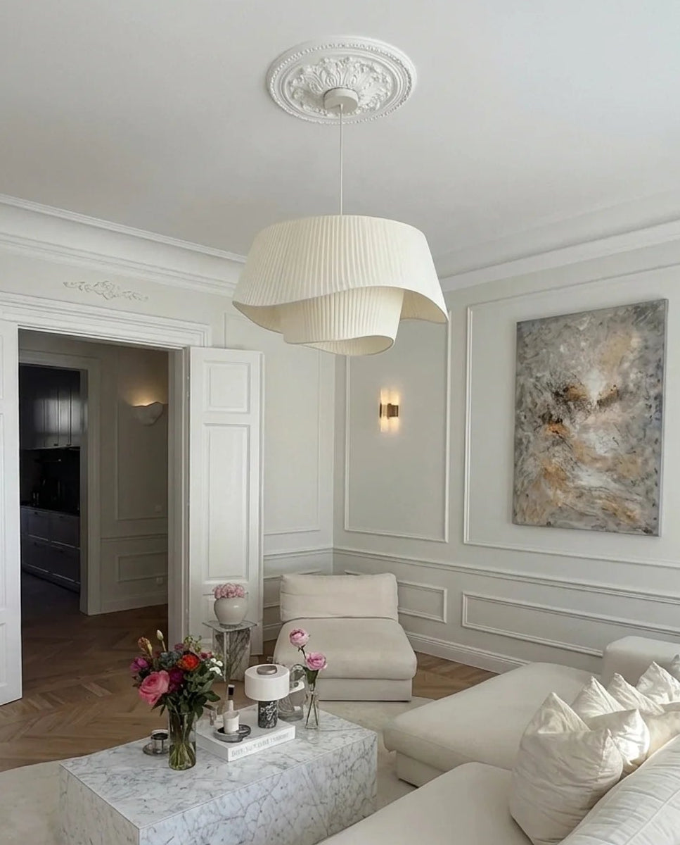 A bright, elegant living room featuring a cream-colored sectional sofa, a matching armchair, a marble coffee table, and a large cream pleated pendant light hanging from a decorative ceiling medallion.