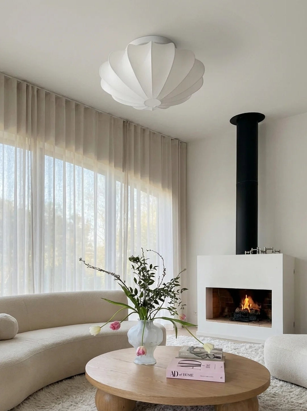 A modern living room featuring a curved beige sofa, a round wooden coffee table with a vase of flowers, a white shag rug, a black chimney pipe, and a white fireplace.