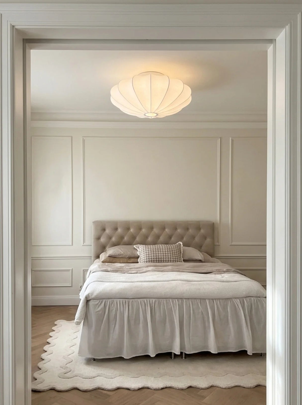 A bedroom scene viewed through a doorway, featuring a bed with white bedding, a tufted beige headboard, a white scalloped rug, and a glowing ceiling light.
