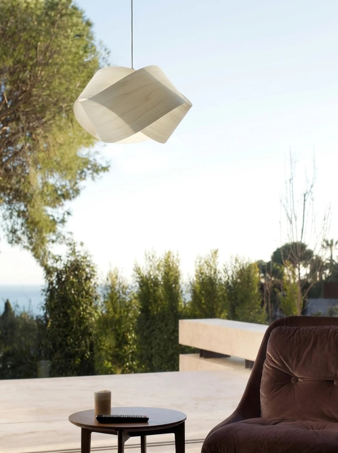 A modern pendant light with a faceted, geometric white shade hangs over a minimalist outdoor patio scene featuring a brown velvet armchair, a small round side table, and a scenic background of trees and the sea.