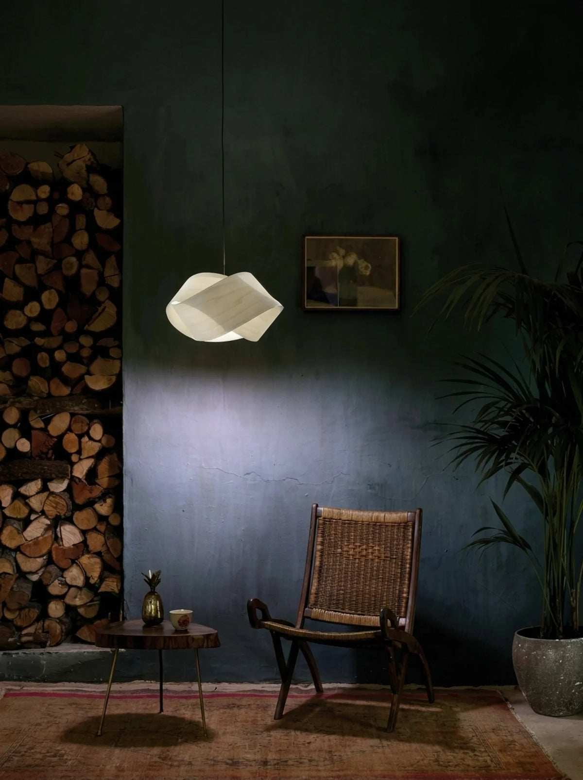 A modern pendant light with a white, organic, cloud-like geometric shade hangs in a dimly lit room with a dark green wall. The scene includes a woven wooden armchair, a small round side table with decorative items, a stack of firewood on the left, and a potted palm on the right.