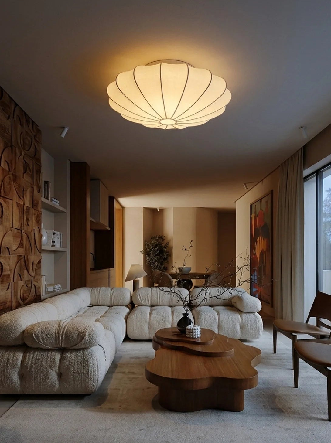 A modern living room featuring a large beige textured sectional sofa, a unique wavy wooden coffee table, mid-century style wooden chairs, and a distinctive scalloped ceiling light.