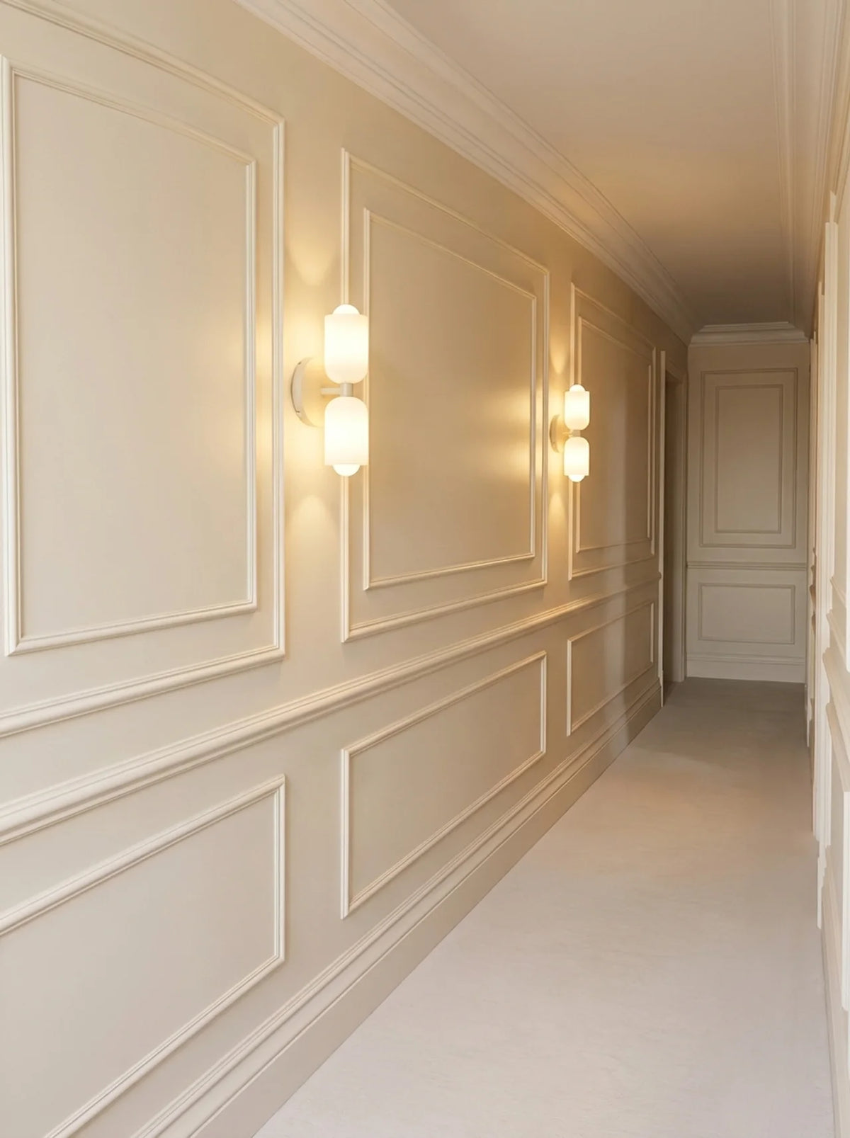 A long hallway with cream paneled walls, decorative molding, and two illuminated wall sconces mounted on the left side. The floor and ceiling are also in a warm off-white tone.