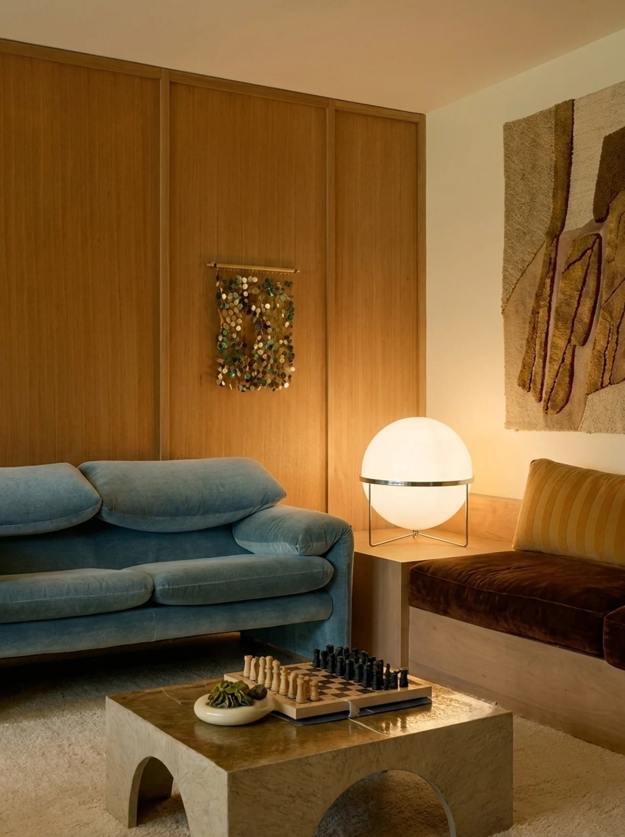 A modern living room interior featuring a blue velvet sofa, a low concrete coffee table with a chess set, a glowing white globe lamp, and warm wood-paneled walls.