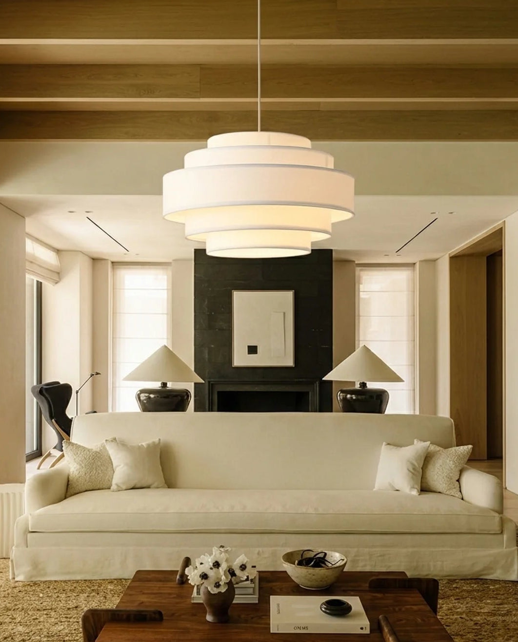 A modern multi-tiered white pendant light hangs in a spacious, warm-toned living room with a cream sofa, a dark wood fireplace, and a wooden coffee table.