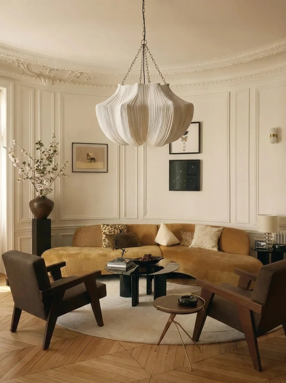 A styled living room interior featuring a curved mustard yellow sofa, two dark brown armchairs, a round black coffee table, a small wooden side table, and a large white pleated pendant light hanging from a chain in a cream-colored room with paneled walls and wood parquet flooring.