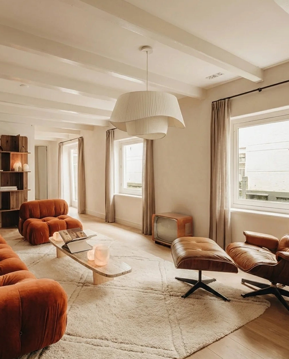 A warm, modern living room featuring a rust-orange velvet sectional sofa, matching lounge chair, and ottoman on a large cream shag rug. The room has exposed white ceiling beams, light wood floors, and large windows with beige curtains.
