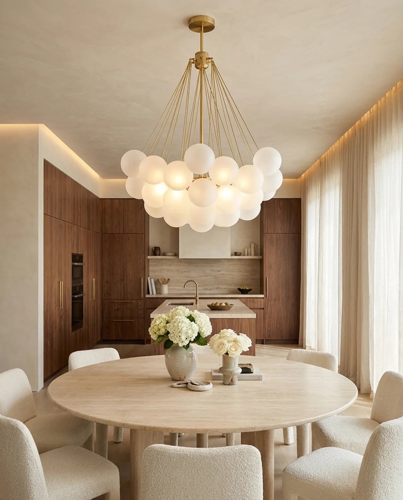A modern dining room featuring a round light wood table surrounded by cream upholstered chairs, with a large multi-light bubble chandelier hanging above. In the background is a warm wood kitchen with built-in lighting and sheer curtains.
