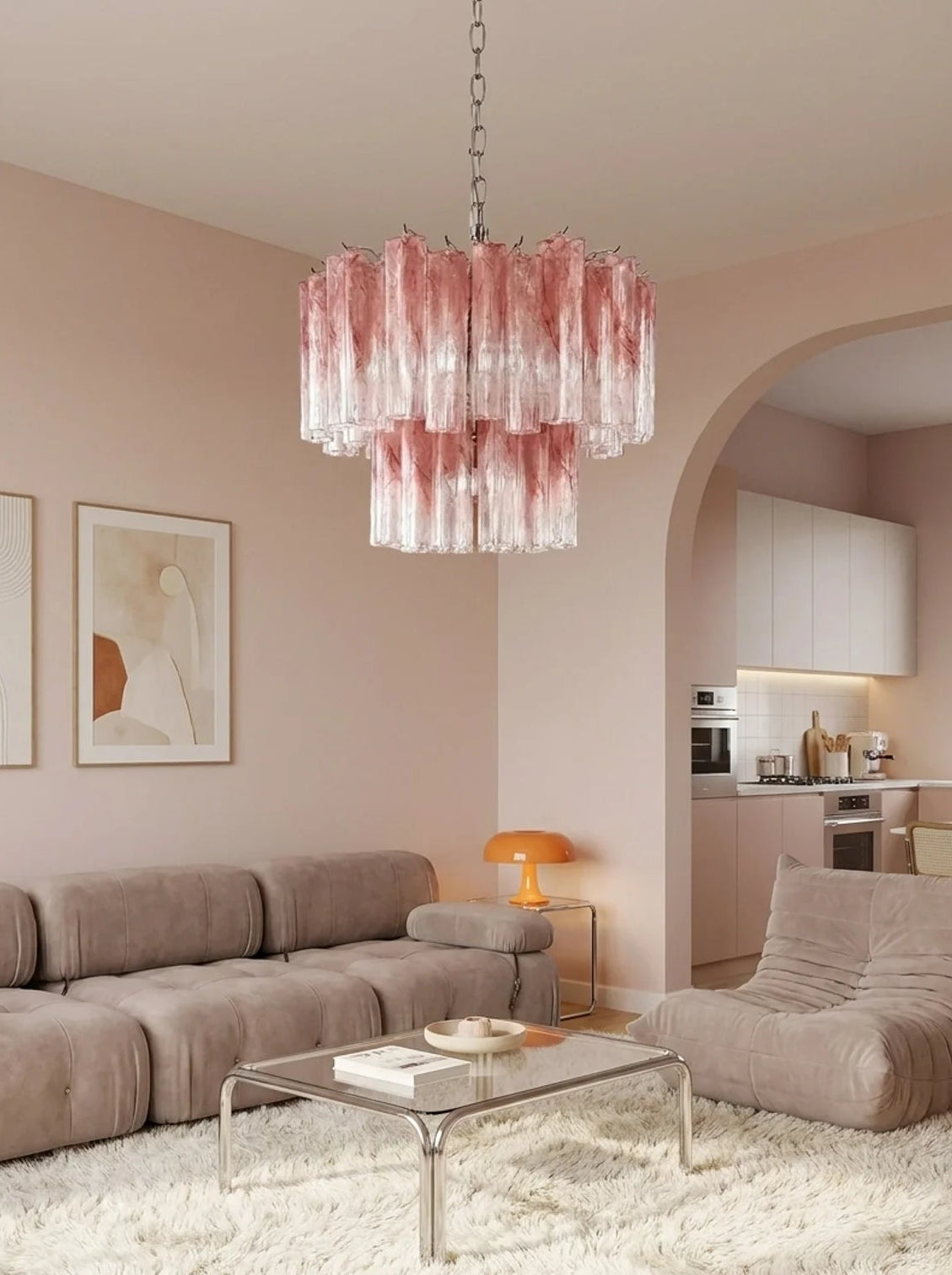 A modern living room featuring a pink and clear textured glass chandelier hanging above a beige sectional sofa and glass coffee table on a white shag rug. The room has blush pink walls, a built-in kitchen in the background, and a matching beige lounge chair.