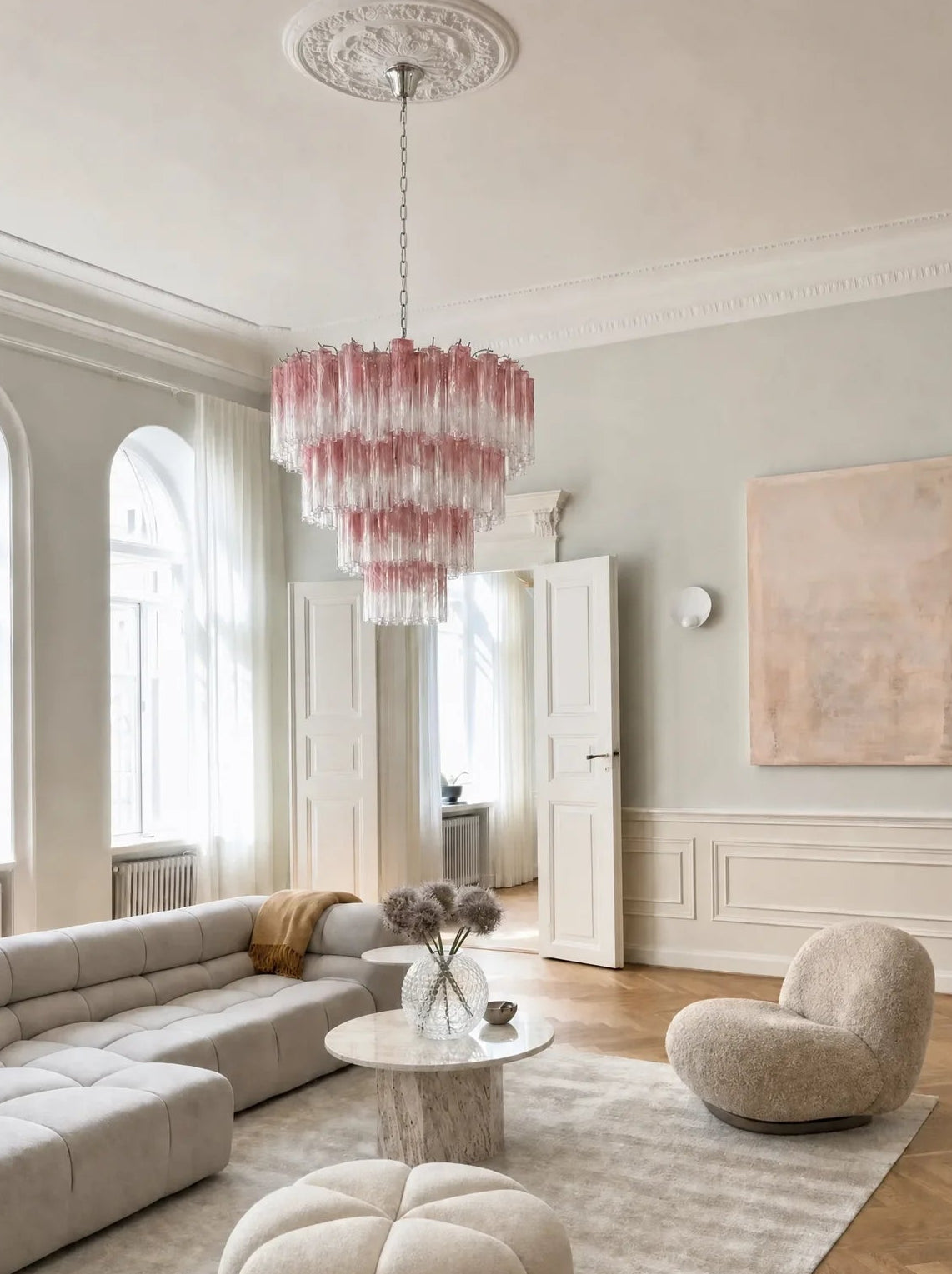 A modern living room featuring a tiered pink glass chandelier hanging from a decorative ceiling medallion, above a light beige sectional sofa, a round tufted ottoman, and a textured beige accent chair on a light rug.