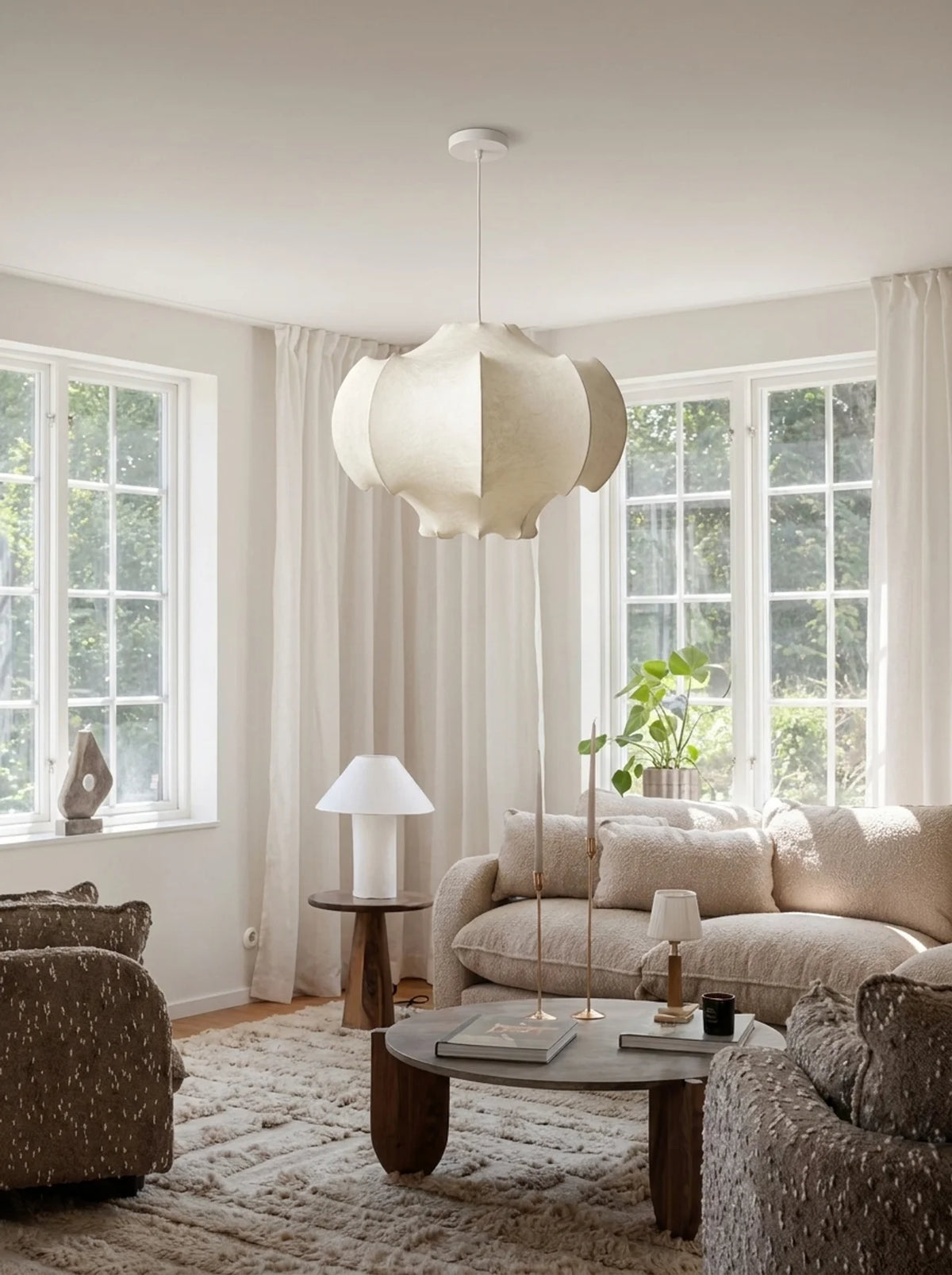 A modern, sculptural white pendant light with a layered, petal-like design hangs in a bright, neutral-toned living room with a beige sofa, wooden coffee table, and large windows.