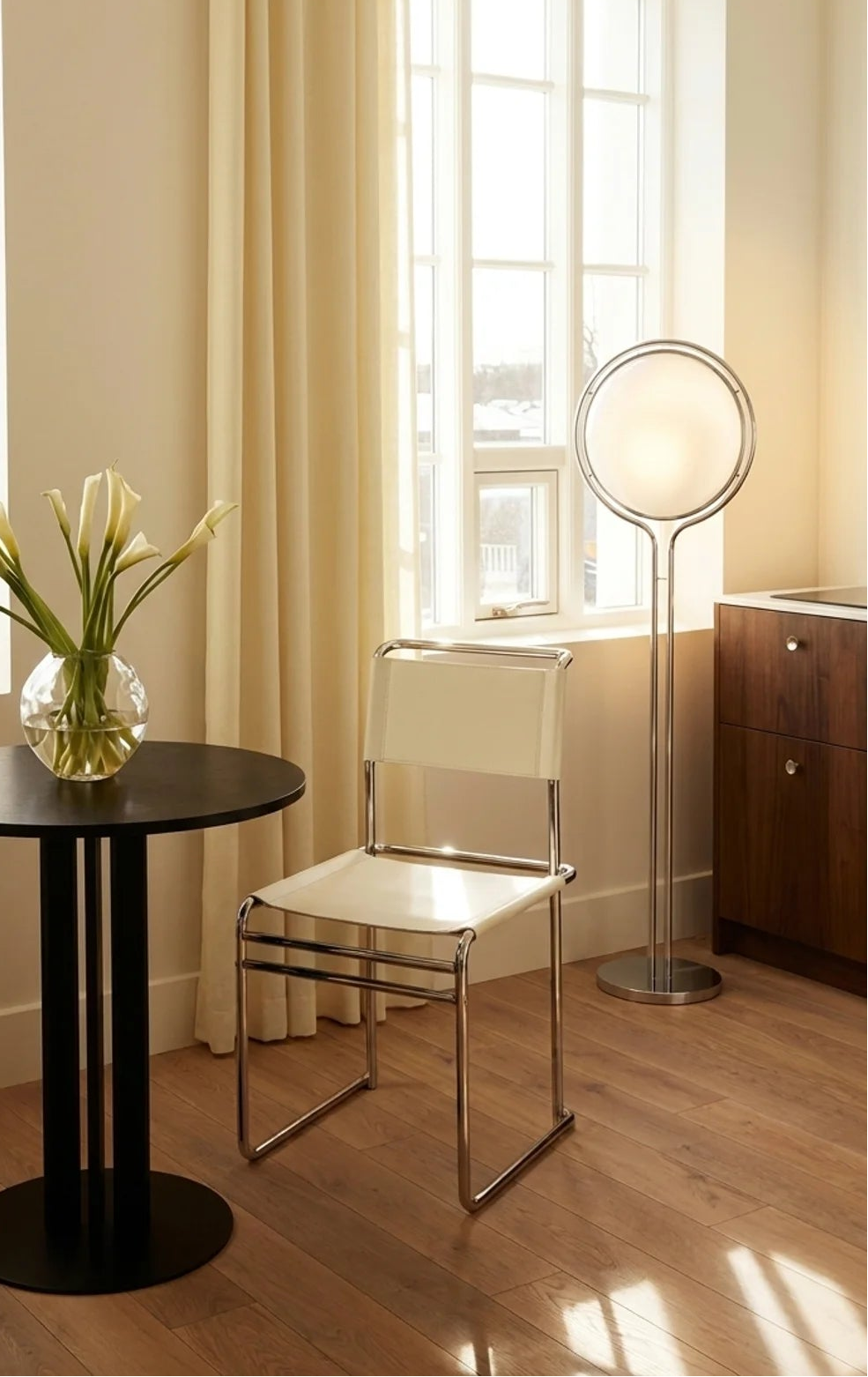 A modern interior scene with a round black side table holding a glass vase of white lilies, two white-and-silver chairs, cream curtains, wood flooring, and a floor lamp near a window.