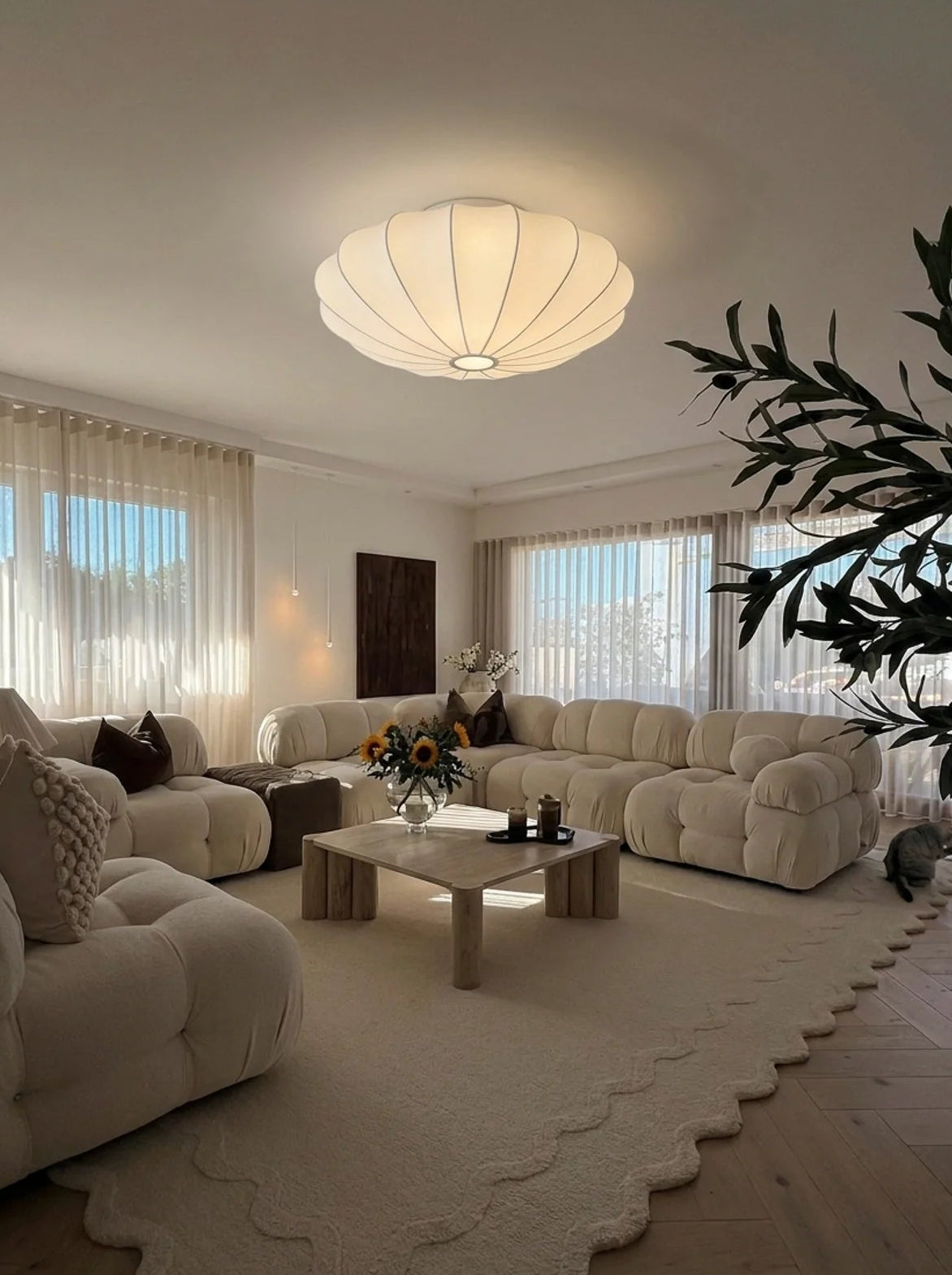 A modern living room featuring a large, sculptural cream boucle sectional sofa, a matching chaise lounge, a scalloped cream area rug, and a central wooden coffee table under a glowing, scalloped ceiling light.