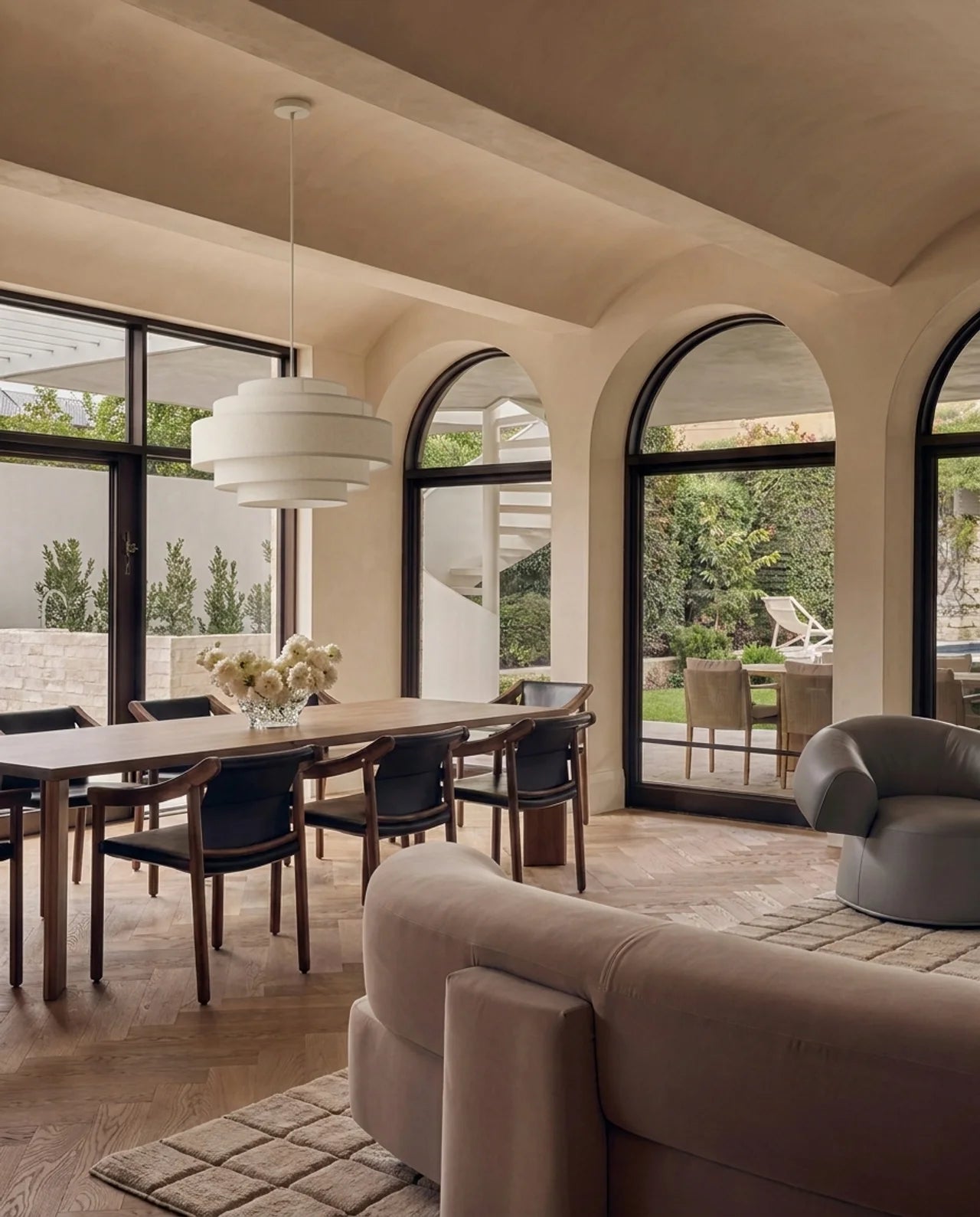 A modern dining room with a rectangular wooden table, six dark upholstered chairs, a white tiered pendant light, and large arched windows looking out to a garden. The room has warm beige walls, wood flooring, and a matching beige sofa in the foreground.