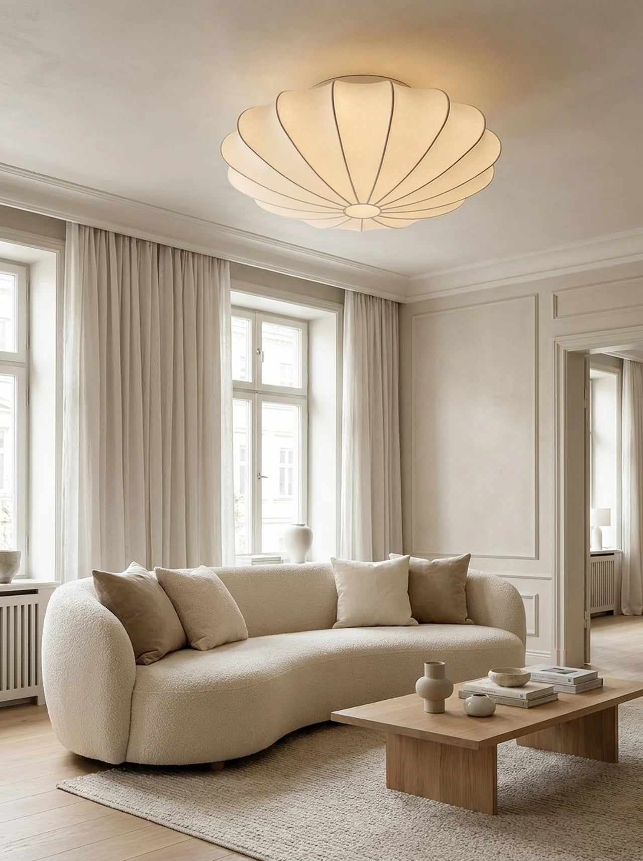 A modern curved beige sofa in a bright, minimalist living room with a matching beige rug, a wooden coffee table, and a distinctive scalloped ceiling light fixture.