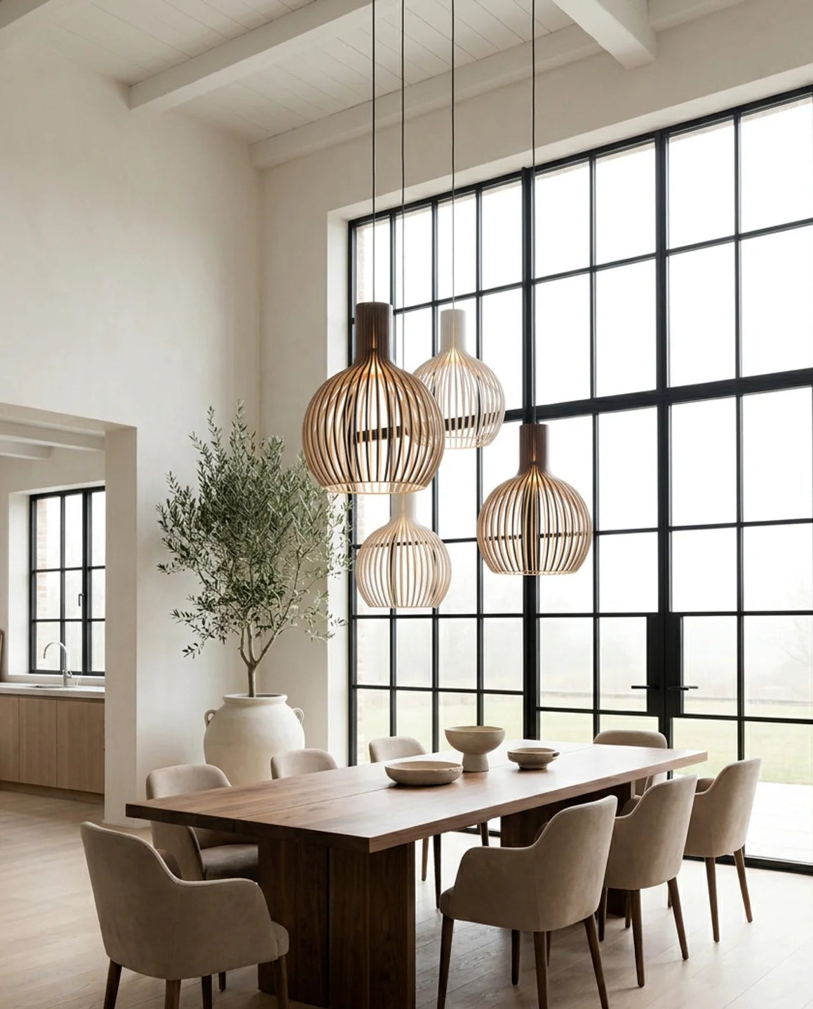 A modern dining room with a long wooden table, beige upholstered chairs, and three woven pendant light shades hanging above. Large black-framed windows dominate the right wall, and a tall potted tree stands near the table.