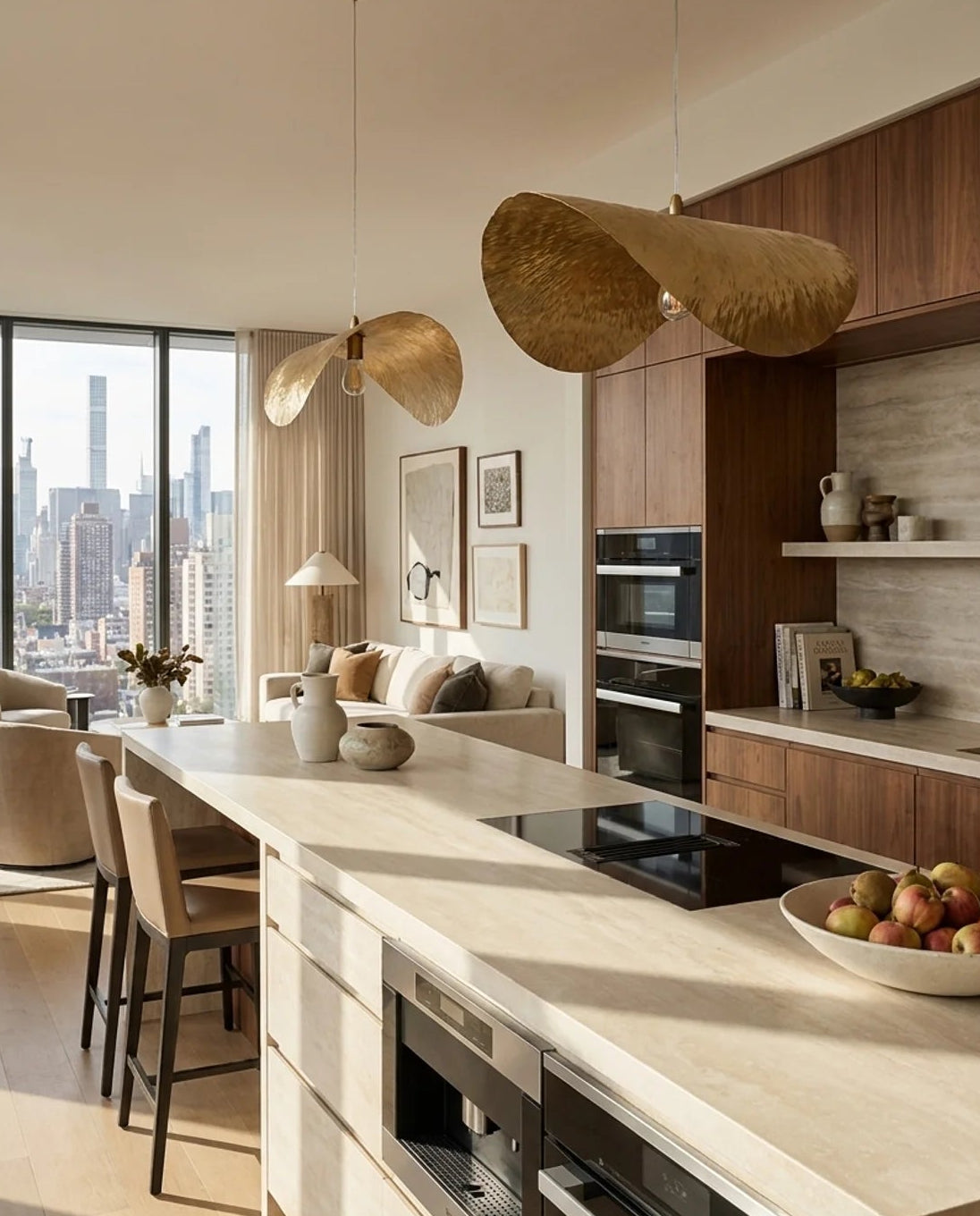 A modern kitchen island with light wood countertops and cabinetry, black built-in appliances, and two large woven pendant lights hanging above. The space is bright with a city skyline visible through large windows, and a bowl of fruit sits on the counter.