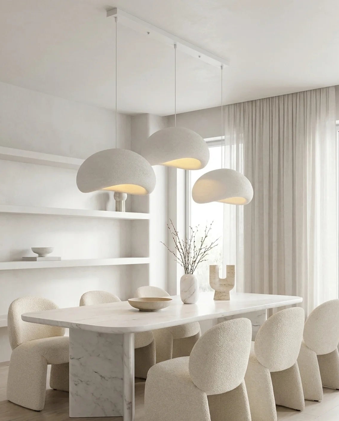 A modern dining room featuring a white marble-topped rectangular table surrounded by textured cream chairs, under three matching dome-shaped pendant lights. The room has light wood floors, built-in white shelves, and sheer curtains.