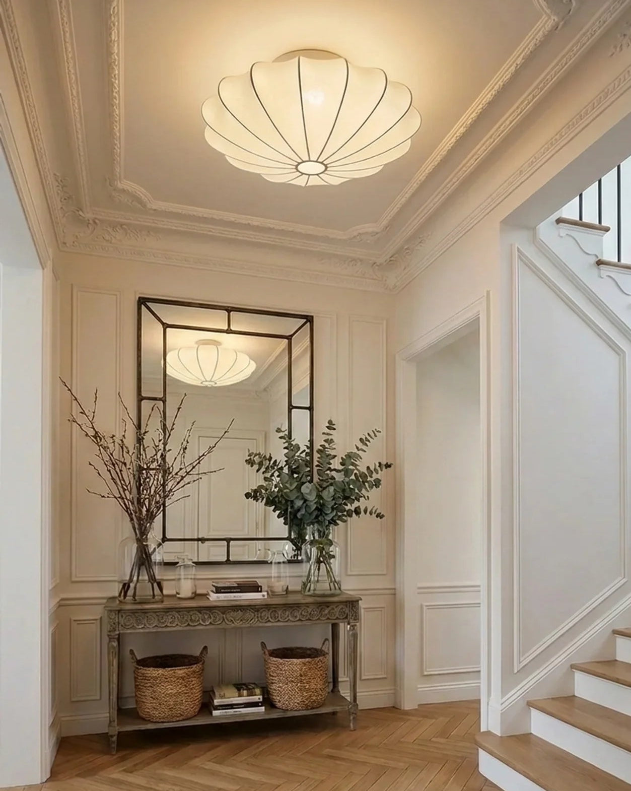 A modern shell-shaped ceiling light fixture with a warm white glass shade and black metal frame, mounted on a decorative white ceiling in an elegant hallway with a console table and mirror.