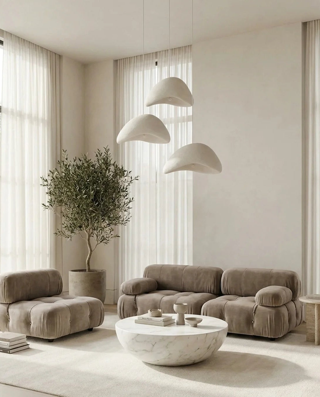 A modern living room featuring a taupe velvet three-seater sofa, a matching armchair, a round white marble coffee table, and a large white area rug. The room has a minimalist design with beige walls, sheer white curtains, three cloud-shaped pendant lights, and a potted olive tree.
